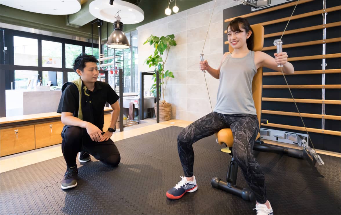 女性がフィットネスマシンでトレーニングをしているところを、男性のトレーナーが見守っている。
