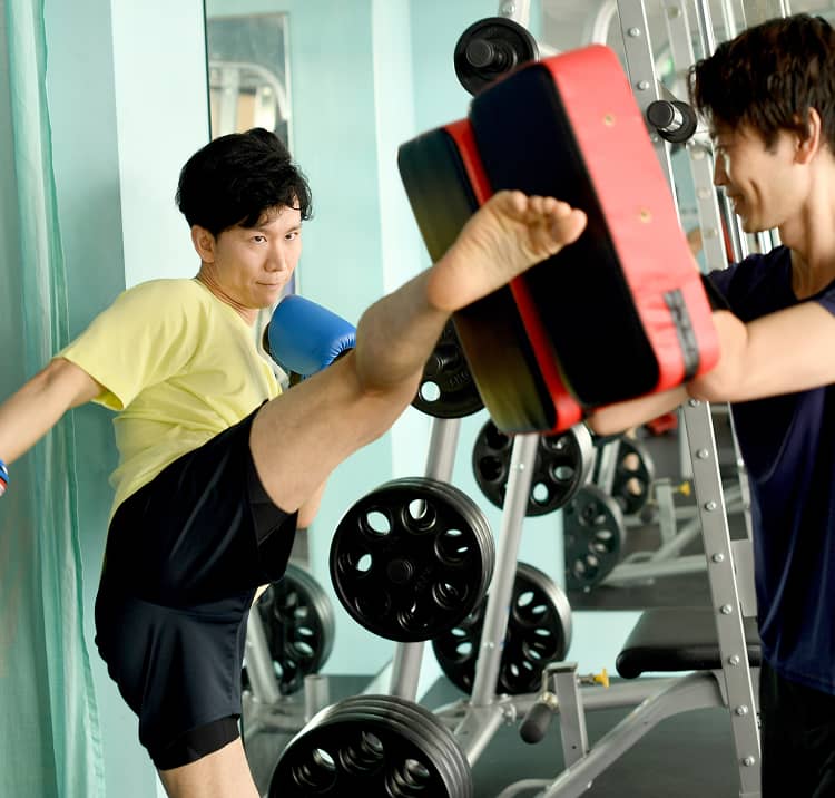 ボクシンググローブをつけた男性がトレーナーのミットを蹴るシーン。