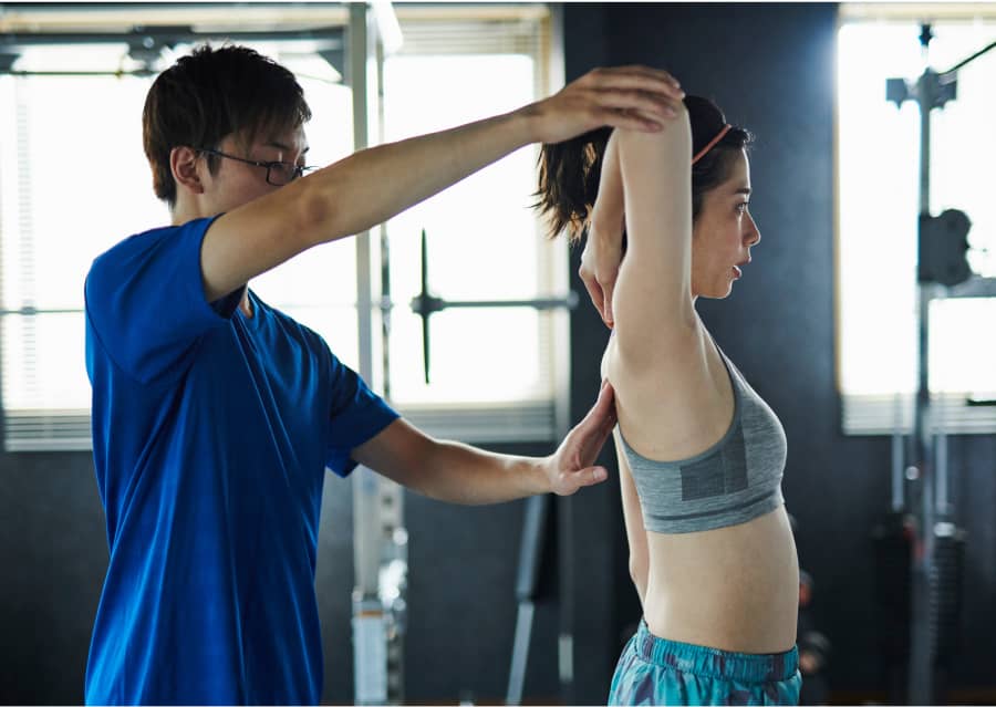 レーナーが女性に腕のストレッチ方法を教えているシーン。女性は腕を上げ、トレーナーが正しいポジションに手を添えている。