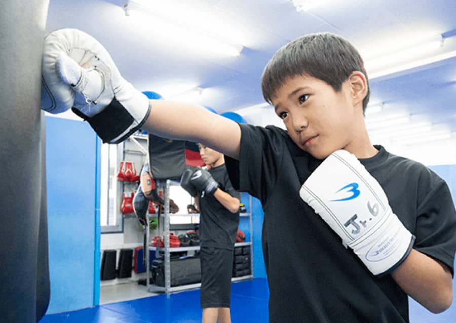 キックボクシングのトレーニングをしている少年が、サンドバッグにパンチをしている様子。白いグローブと集中した表情が特徴。